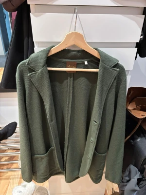 Olivgrön kavaj från Stenströms - Stilren olivgrön kavaj från Stenströms i stickad struktur, 100% merino ull. Kavajen har två framfickor, klassisk krage och knappar framtill. Perfekt för en avslappnad men ändå uppklädd look. Materialet är mjukt och följsamt.