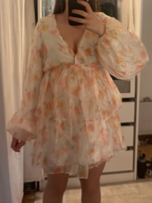 Blommig kortklänning med volang - Söt kortklänning med volanger och v-ringning. Klänningen har ett blommigt mönster i ljusa toner av rosa, orange och vitt samt långa, vida ärmar. Perfekt för sommarens alla tillfällen.