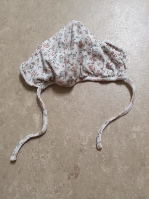 Mössa - Söt mössa från Newbie i mjuk bomull med vitt tyg och små blommor i rosa och grönt. Mössan har knytband under hakan och en klassisk form som sitter bra på huvudet. Perfekt accessoar för en gullig och romantisk look.