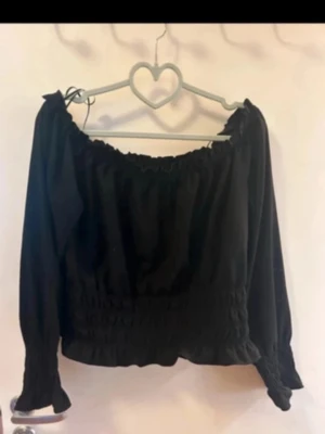 Svart offshoulder blus med smock - Trendig svart blus med offshoulder-design och smockad midja. Långa ärmar med volangdetaljer och elastiska muddar. Perfekt för dig som gillar en romantisk och cool stil.