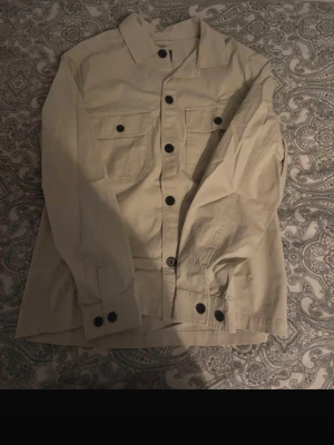 Beige overshirt med svarta knappar - Stilren beige overshirt med svarta knappar och två stora bröstfickor. Skjortan har klassisk krage och lång ärm med knapp vid manschetten. Perfekt att bära öppen eller stängd för en avslappnad look.