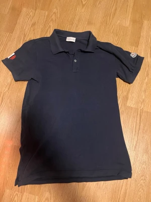 Mörkblå pikétröja från Moncler - Klassisk mörkblå pikétröja från Moncler med krage och knappar framtill. Tröjan har korta ärmar och Moncler-logga på ena ärmen. Perfekt för en clean och stilren look. Storlek 16y som motsvarar S/xs. Bra men använt skick😊