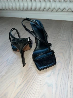 Svarta sandaler med klack och remmar - Säljer ett par svarta pumps från Bianco med smala remmar och fyrkantig tå. Skorna har en hög klack och en snygg ormmönstrad yta i konstläder. Perfekta för dig som vill ha en trendig och elegant look. 