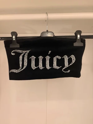 Svart Juicy Couture tubtopp med strass - Säljer en svart tubtopp från Juicy Couture med glittrande strassbokstäver som stavar 'Juicy' framtill. Perfekt för en trendig och edgy look. Materialet känns mjukt och stretchigt, vilket gör den bekväm att bära. 🌟🌟