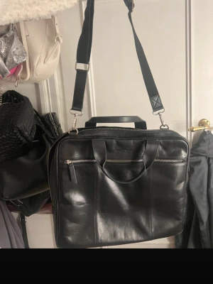 Svart datorväska i läder - Snygg svart datorväska i äkta läder med både handtag och justerbar axelrem. Väskan har en stor ytterficka med dragkedja och stilren design, perfekt för laptop och viktiga prylar. Passar dig som vill ha en klassisk och stilren look till skolan eller jobbet.