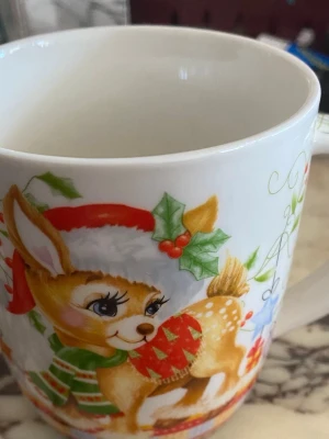 Julmugg med ren och dekorationer - Säljer en vit mugg med färgglatt julmotiv. Muggen har en söt ren med tomteluva och halsduk, samt julgranskulor, stjärnor och mistel runt om. Perfekt för varma drycker under vintern och julen. Tillverkad i keramik.