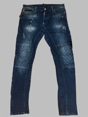 DSQUARED2 Jeans - DSQUARED2 blåa jeans med faded tvätt, slitna detaljer och quiltade paneler vid knäna. Modellen har även en dekorativ dragkedja vid framfickan.