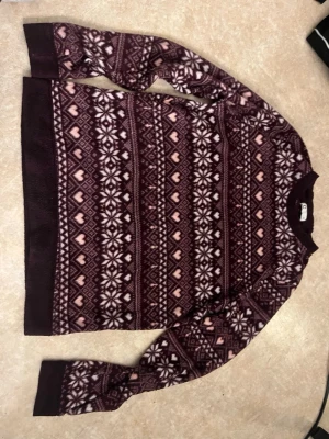 Vinröd mönstrad stickad tröja - Fleece tröja i vinrött med vita och rosa hjärtan och snöflingemönster. Rund halsringning och långa ärmar. Ribbstickade muddar vid ärmslut och nederkant. Perfekt för kalla dagar och mysiga stunder.