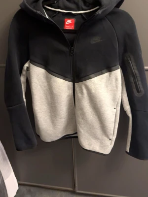 Nike tech hoddie  - Storlek barn xlNike hoodie i svart och grått med dragkedja framtill och huva. Stort Nike-logga på bröstet och en praktisk ficka med dragkedja på ena ärmen. Tillverkad i mjukt sweatshirtmaterial, perfekt för en avslappnad streetwear-look.
