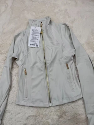 Ljusgrå träningsjacka från Lululemon - Snygg ljusgrå träningsjacka från Lululemon med guldfärgade dragkedjor och hög krage. Jackan har figurnära passform, långa ärmar och två fickor med dragkedja framtill. Materialet är stretchigt och passar perfekt för sport och träning.
