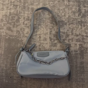 Ljusblå axelväska med kedjedetalj - Trendig ljusblå handväska. Väskan har en justerbar axelrem och en silverfärgad kedjedetalj framtill. Enkel, rektangulär form och diskret logga på framsidan. Perfekt för dig som gillar stilrena accessoarer.