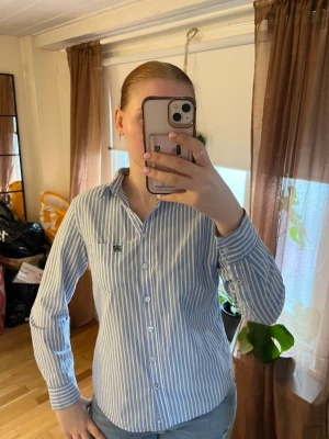 Blåvit randig skjorta från Hampton Republic - Snygg blå och vit randig skjorta från Hampton Republicmed klassisk krage och knäppning framtill. Skjortan har lång ärm och en liten broderad logga på bröstfickan. Perfekt för en stilren och fräsch look.