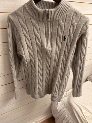 Grå kabelstickad tröja Polo Ralph Lauren - Kabelstickad grå tröja från Polo Ralph Lauren med halv dragkedja och ribbade muddar. Tröjan har det klassiska svarta Polo-logotypen broderad på bröstet och är gjord i mjukt material. Storleken är L men längden är 63cm så passar mer storleken S/M