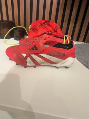 Adidas Predator röda fotbollsskor - Säljer ett par röda Adidas Predator fotbollsskor med svarta och vita detaljer. Skorna har klassisk låg profil, snörning och dobbar för spel på gräs. Tillverkade i syntetmaterial och mesh för bra ventilation. Medföljer matchande röd påse med Predator-tryck.