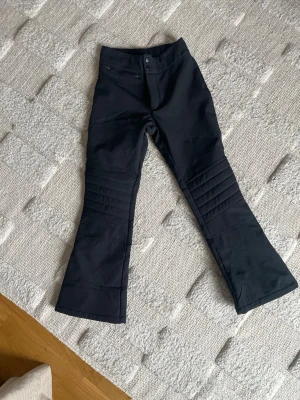 Skidbyxor svarta! - Säljer ett par svarta täckbyxor med utsvängda ben och quiltade detaljer över knäna. Byxorna har knapp och dragkedja framtill samt en ficka med dragkedja. Perfekta för kalla dagar när du vill hålla dig varm och snygg. Från lager 157. Använda en gång. Storlek 150. 