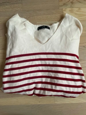 Vit tröja med röda ränder från Mango  - Vit långärmad tröja från Mango med röda horisontella ränder över bröstet och ärmarna. Tröjan har v-ringning och är gjord i ett tunt, mjukt material som känns skönt mot huden. Enkel och stilren design som passar till många looks. Fin att ha över en skjorta. 