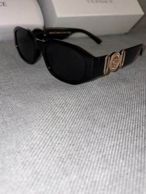 Svarta solglasögon från Versace - Snygga svarta solglasögon från Versace med rektangulära bågar och ikoniska guldfärgade Medusa-detaljer på sidorna. Glasögonen har en chunky design och ger en lyxig vibe till din outfit. Perfekta för dig som vill sticka ut med en klassisk accessoar. Tveka inte på att höra av vid funderingar!
