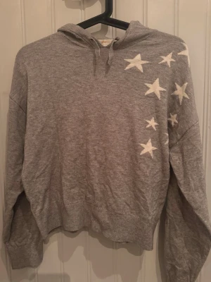 Grå stickad hoodie med stjärnor - Grå stickad hoodie med vita stjärnor på axeln och ryggen. Tröjan har huva med dragsko, långa ärmar och ribbade muddar. Perfekt för en avslappnad och trendig look. Materialet ser mjukt och bekvämt ut.