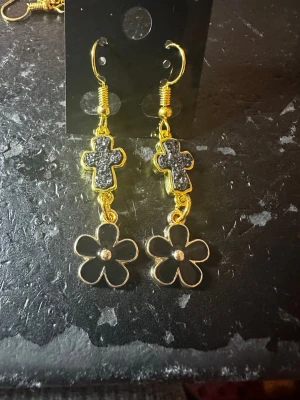 Svart-guldiga örhängen med kors & blomma - Unika örhängen i guldmetall med svarta detaljer. Hängande design med glittrande kors och blommor längst ner. Perfekta för dig som vill sticka ut med coola accessoarer.