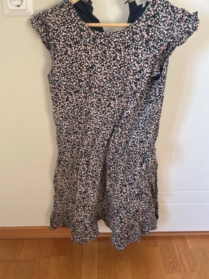 Blommig kortklänning i blått och vitt - Söt kortklänning med blommigt mönster i blått, vitt och rosa. Klänningen har rund hals, korta ärmar och lätt utställd kjol. Perfekt för varma dagar och ger en fräsch och ungdomlig look. Tillverkad i ett mjukt och lätt material som känns skönt mot huden.