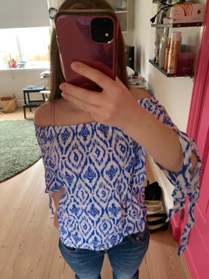 Blåvit offshoulder blus med knyt - Snygg blus i blått och vitt med grafiskt mönster. Offshoulder-modell med knytband på ärmarna och lös passform. Perfekt till jeans eller kjol för en fräsch look.