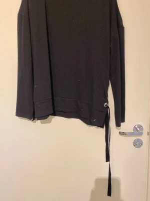 Svart tröja snörning i sidan - Cool svart tröja med unika snörningsdetaljer i sidan och stora metallöljetter. Oversized passform med långa ärmar och ribbade muddar. Tillverkad i mjukt sweatshirtmaterial som känns skönt mot huden. Perfekt för en avslappnad streetwear-look.