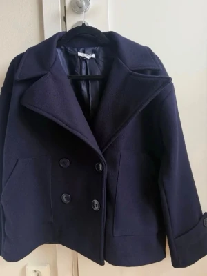 Mörkblå kappa - Stilren mörkblå kappa med bred krage och dubbelknäppning framtill. Jackan har två framfickor och är tillverkad i ett tjockare tyg som ger en klassisk look. Original pris 757kr💕