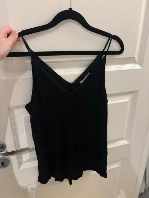 Svart linnetopp med smala axelband - En enkel och stilren svart linnetopp med smala axelband och v-ringad hals både fram och bak. Toppen har en lös passform och är perfekt att matcha med jeans eller kjol. Materialet känns mjukt och lätt mot huden.