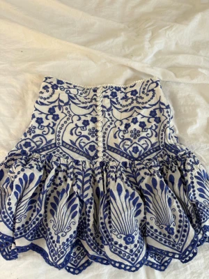 Vit och blå broderad kortkjol - Supersöt kortkjol i vitt med blått broderat mönster och dekorativa knappar framtill. Kjolen har volangkant och ett unikt, detaljerat blommönster som ger en bohemisk vibe. Perfekt för dig som vill sticka ut med en feminin och trendig look.