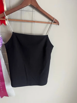 Svart linnetopp med smala axelband - Snygg svart linnetopp med tunna axelband i silverfärg. Enkel och stilren design som passar perfekt till jeans eller kjol. Toppen har rak skärning och ärmlös modell för en clean look.