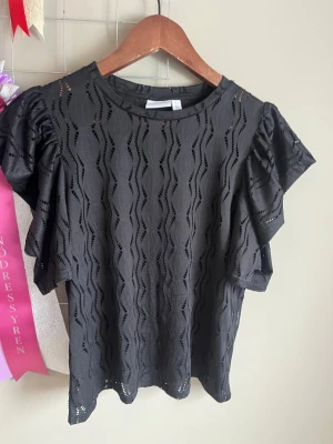 Svart blus med volangärmar - Säljer en svart blus med vågigt hålmönster och volangdetaljer på ärmarna. Blusen har rund halsringning och är tillverkad i ett lätt, luftigt material som passar perfekt till sommaren. Superfin att styla med både jeans och kjol!