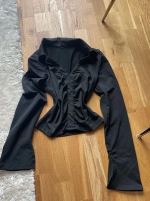 Svart croppad v-ringad topp - Svart croppad topp med v-ringning och långa ärmar. Toppen har en rynkad detalj framtill. Korta slits vid kanten av ärmarna. 