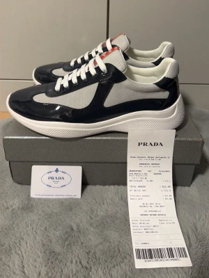 Prada cups  - Snygga marinblå och vita Prada-sneakers med kvitto och autenticitetkort. De har vita skosnören och röda detaljer på plösen. Ovandelen är gjord av en mix av glansigt material och mesh av hög kvalitet, vilket ger en modern och sportig look. Den tjocka vita sulan ger extra komfort och stil.  Priset kan diskuteras, så tveka inte att höra av dig!