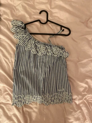 Randig oneshoulder topp - Säljer en blåvit randig oneshoulder topp med volang och broderad spetskant. Toppen har smal axelrem och ärmlös design, perfekt för sommarens varma dagar. Materialet känns lätt och luftigt, och spetsdetaljerna ger en söt touch. Är storlek 152 fast skulle säga att den passar som en XS.
