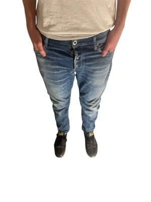 Blåa G-Star Raw jeans herr - Säljer ett par blåa jeans från G-Star Raw med avslappnad passform och tydliga slitningar framtill. Jeansen har knappgylf, klassiska fem fickor och en brun läderpatch med logga bak i midjan. Perfekta för en casual streetstil.