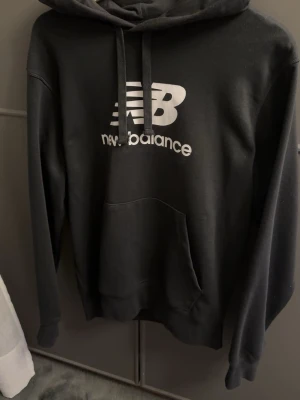 Svart hoodie från New Balance - Säljer en svart hoodie från New Balance med stor vit logga och text på bröstet. Hoodien har huva med dragsko och en stor magficka framtill. Tillverkad i mjukt material som passar perfekt för en avslappnad stil.