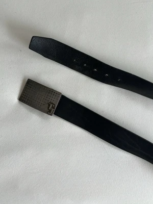 Svart skärp med metallspänne från Tiger of sweden  - Stilrent svart skärp i skinn med ett rektangulärt metallspänne dekorerat med små hål och TJ-logga. Perfekt accessoar för att lyfta din outfit med en modern touch.