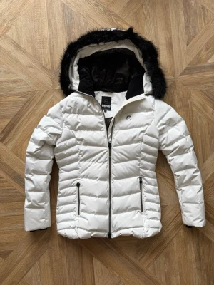 Vit dunjacka från 8848 Altitude - Snygg vit dunjacka från 8848 Altitude med svart foder och huva med svart fuskpäls. Jackan har två dragkedjeförsedda fickor framtill och diskret logga på bröstet. Perfekt för kalla vinterdagar och riktigt varm tack vare dunfyllningen. En svag sminkfläck på bild 2, därav det låga priset.  