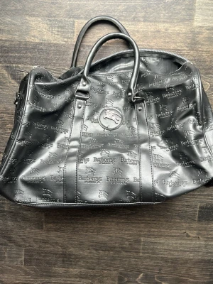 Svart resväska från Burberrys - Cool svart resväska från Burberrys of London i skinn med präglad logotyp och ridmotiv över hela väskan. Väskan har dubbla handtag och en klassisk, rymlig form som passar perfekt när du ska ut och resa. Stängs med dragkedja och har snygga detaljer i metall.