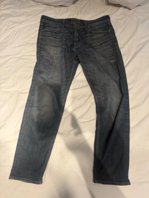 Blå raka jeans med taper - Säljer ett par klassiska blå jeans med raka ben och diskreta slitningar. Jeansen har fem fickor, knappgylf och en läderpatch bak i midjan. Perfekta till en avslappnad stil och tillverkade i denim. Storlek 33/32
