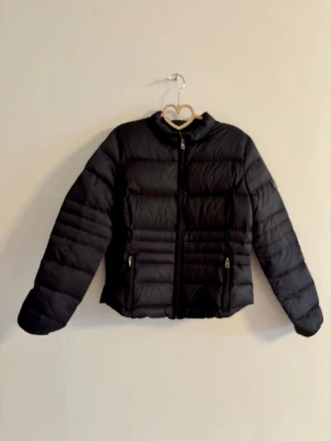 Svart pufferjacka med dragkedja - Snygg svart pufferjacka med raka kanaler och hög krage. Jackan har två framfickor med dragkedja och stängs med en hel dragkedja framtill. Perfekt för kalla dagar och ger en clean look. Materialet är glansigt och quiltat för extra värme.
