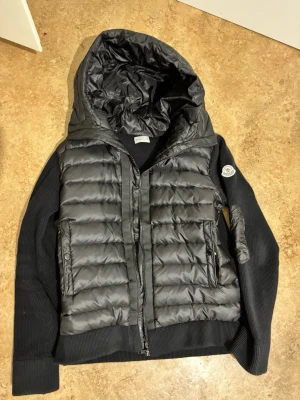 Moncler cardigan - Snygg svart dunväst från Moncler med stickade svarta ärmar och stor huva. Västen har quiltad design, dragkedja framtill och två fickor med dragkedja. Moncler-logga på ärmen ger en exklusiv touch. Perfekt för lager-på-lager och streetstyle.