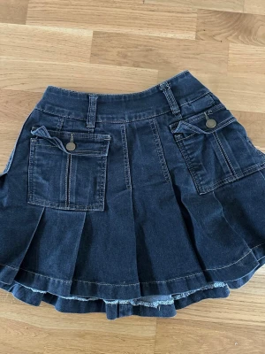 Svart jeanskjol med fickor - Kort svart jeanskjol med två stora fickor framtill och dekorativa knappar. Kjolen har breda veck och bälteshällor i midjan. Tillverkad i denim med synliga sömmar och dragkedja bak. Perfekt för en avslappnad och trendig look!!🤗
