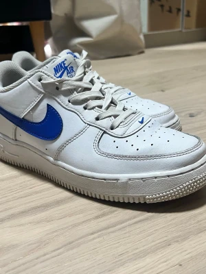 Nike Air Force 1 vita/blå sneakers - Klassiska Nike Air Force 1 sneakers i vitt läder med blå Swoosh, lite använda och tvättas innan leverans 