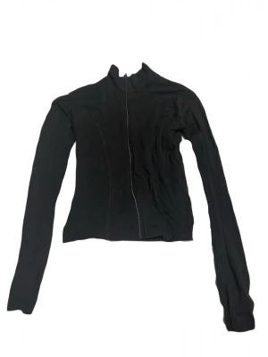 Svart ziptröja med hög krage - Säljer en svart långärmad ziptröja med hög krage och figurnära passform. Tröjan har dragkedja framtill och är perfekt för en stilren och enkel look. Passar till det mesta och är lätt att matcha med andra plagg.