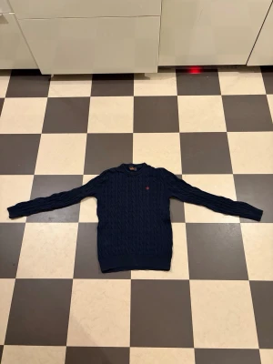Mörkblå kabelstickad tröja Morris - Kabelstickad mörkblå tröja från Morris med rund halsringning och ribbade muddar. Tröjan har ett broderat rött emblem på bröstet och är tillverkad i mjukt material. Perfekt för en stilren och klassisk look. Det står m men passar mer som storlek S