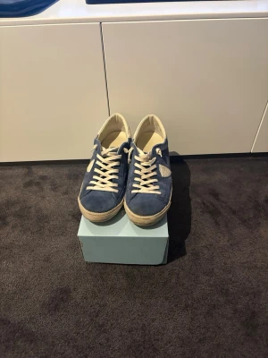 Blåa sneakers från Philippe Model - Snygga blåa sneakers från Philippe Model med ovandel i mocka och vita detaljer. Skorna har vita snören, vit sula och en ikonisk sköldlogga på sidan. Perfekta för en avslappnad och trendig stil.