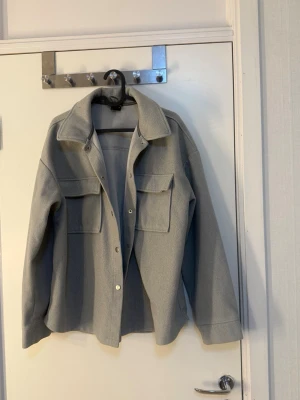 Ljusgrå overshirt med fickor ginatricot  - Stilren ljusgrå overshirt med klassisk krage och två stora bröstfickor med lock. Rak passform och knäppning framtill med tryckknappar. Perfekt lager-på-lager-plagg i ett mjukt, vävt material. 