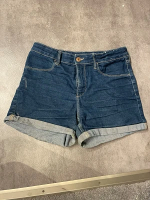 Mörkblå jeansshorts med uppvik - Snygga mörkblå jeansshorts med klassisk femficksmodell och uppvikta benslut. Shortsen har synliga sömmar i kontrastfärg och stängs med knapp och dragkedja. Perfekta för sommaren och enkla att matcha med olika toppar.