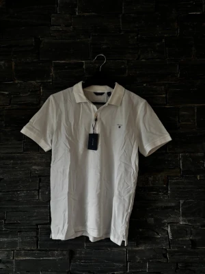 Vit piké t-shirt från Gant - Klassisk vit piké t-shirt från Gant med korta ärmar och krage. Tillverkad i mjuk bomull och har små knappar framtill. Diskret Gant-logga broderad på bröstet. Perfekt för en clean och stilren look.